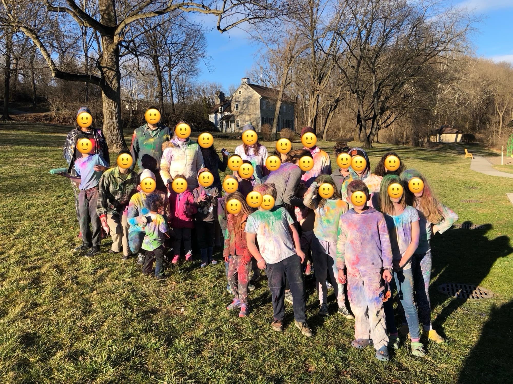 A big group of homeschool families covered in color-run powder on a park lawn, faces covered with smiley emoji for privacy